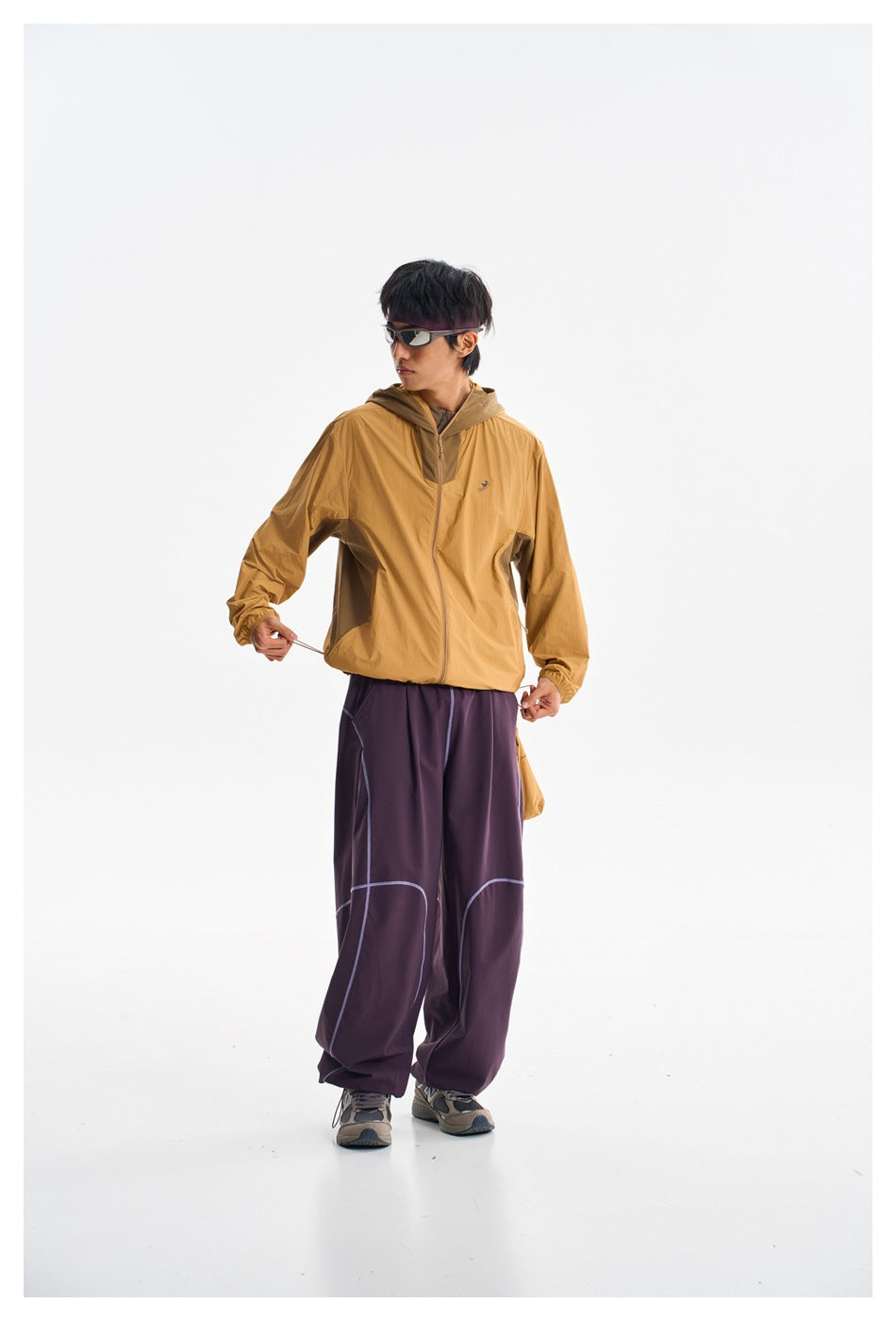 Model wearing a mustard zip-front hooded jacket and loose plum pants with pale seam detailing, shown from the front.