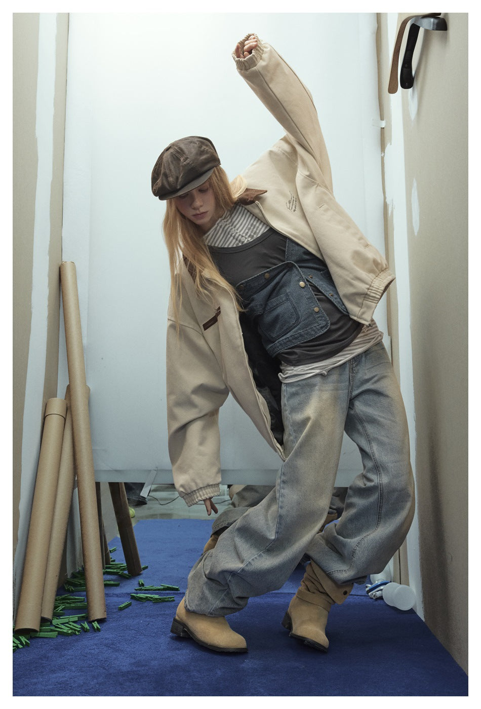 Model poses in a cream zip jacket with brown collar, layered tops, faded wide-leg jeans, and brown cap.