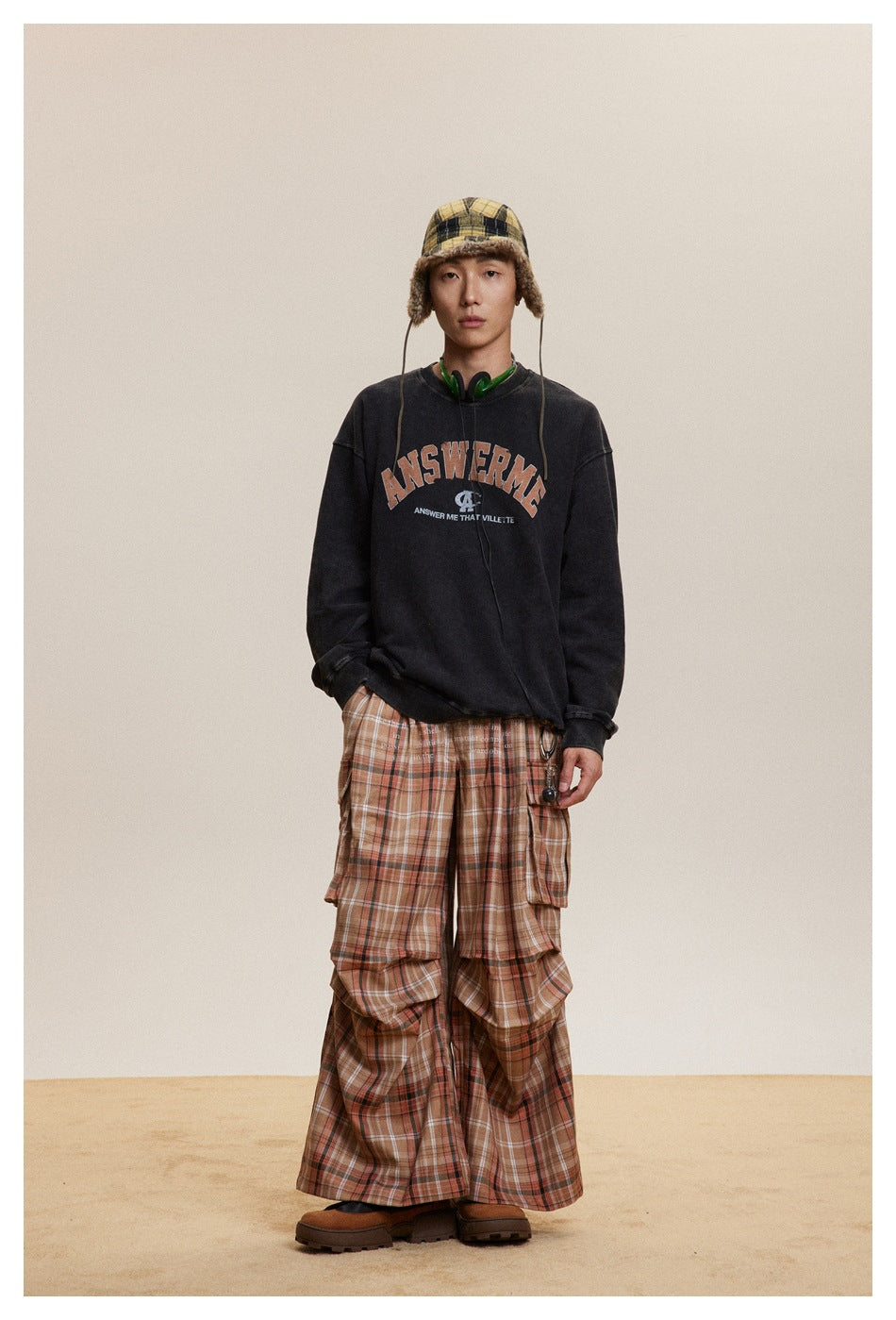 Model wearing black washed crewneck sweatshirt with ANSWERME graphic, plaid cargo pants, fur-trim hat and boots