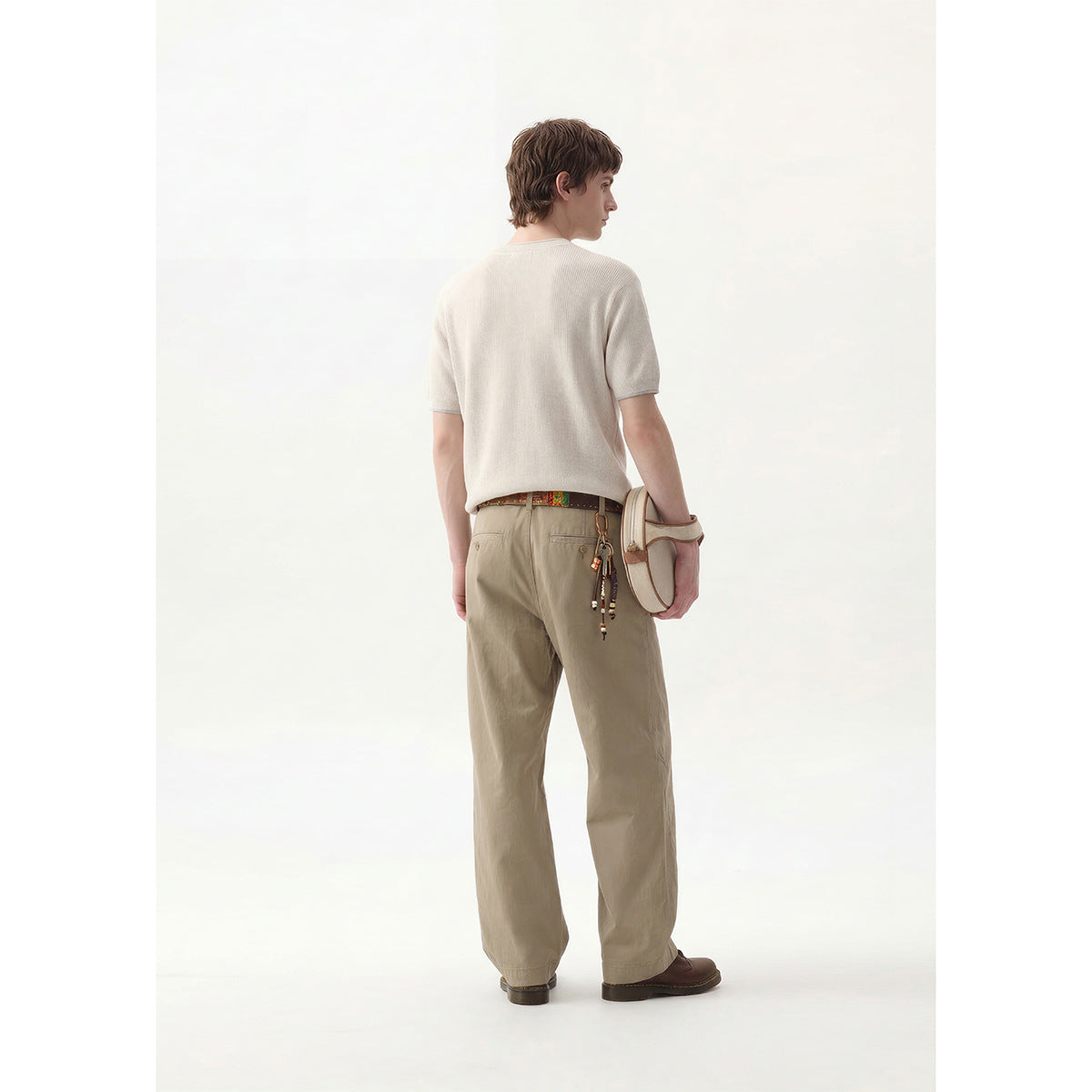 Rear three-quarter view of a model in an off-white short-sleeve top and khaki trousers, carrying a cream shoulder bag.