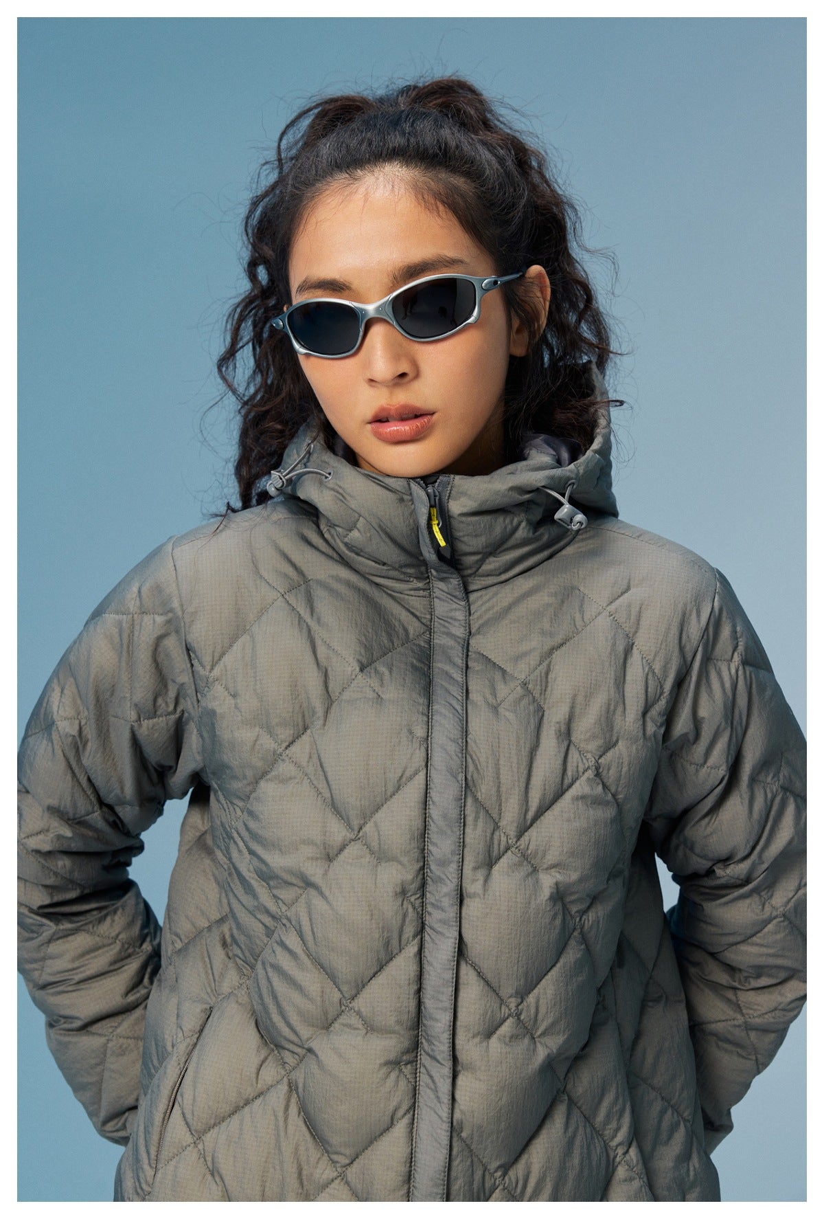 Front view of a model in a gray diamond-quilted hooded coat with a high collar and front closure.