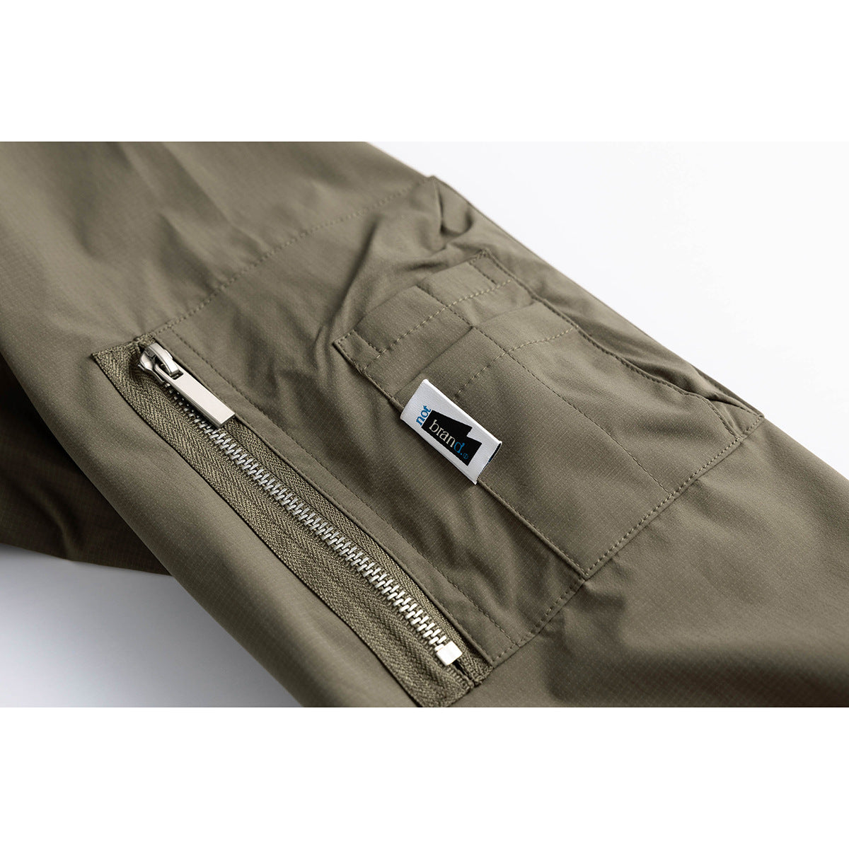 Close-up of an olive jacket's zippered sleeve pocket and stitched patch pocket with a small sewn label.