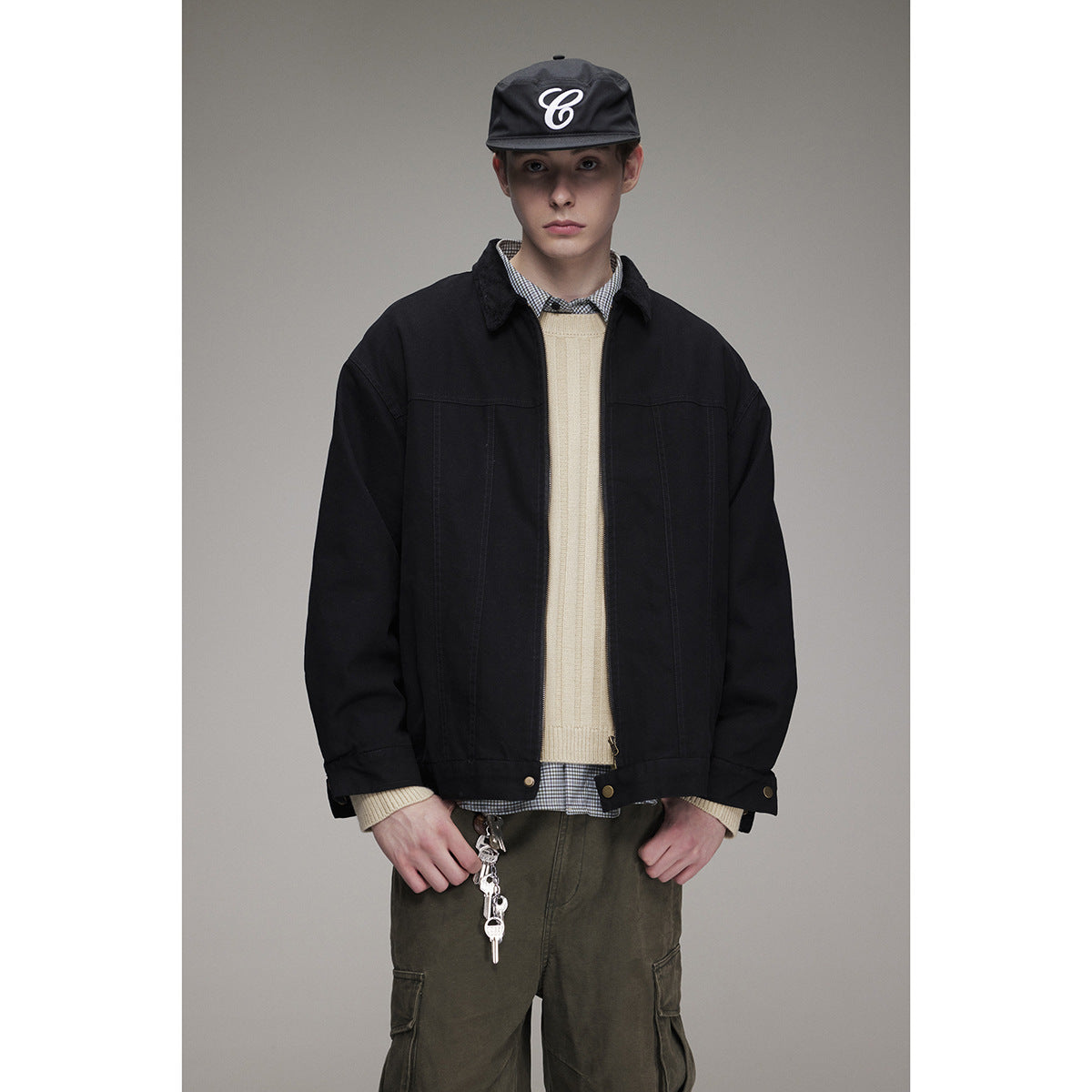 Model wearing a black zip-front jacket over a cream sweater and checked shirt, with a black cap and olive cargo pants.