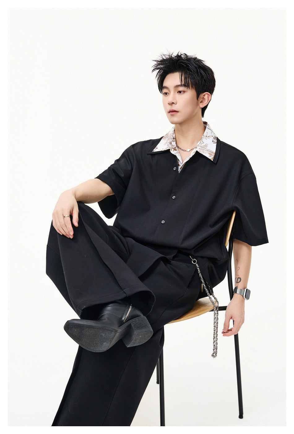 Model seated on a chair wearing a black short-sleeve shirt with floral under-collar and black wide-leg trousers