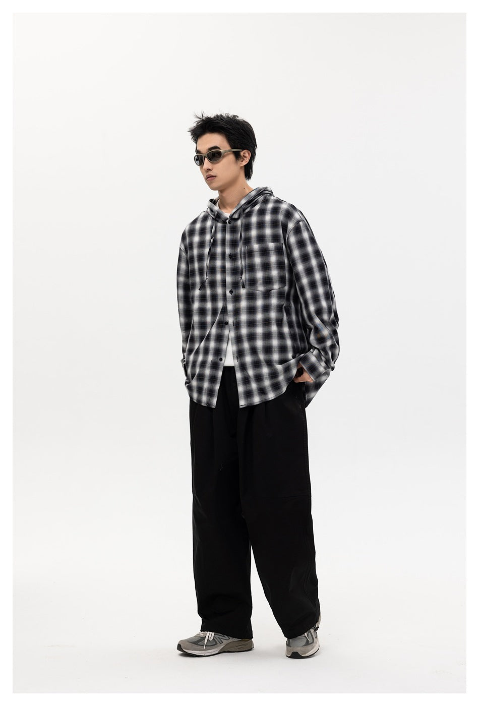 Model wearing black plaid hooded shirt jacket open over a white tee with wide black pants and gray sneakers