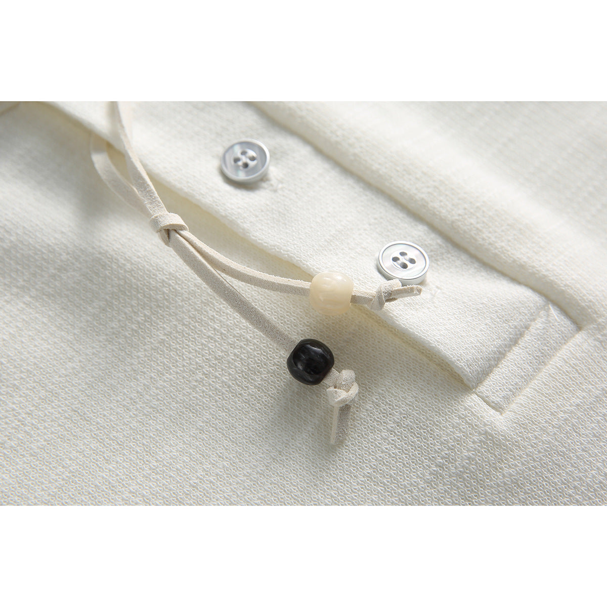 Close-up of an ivory shirt's textured fabric, buttons, and knotted drawcord with contrasting beads.
