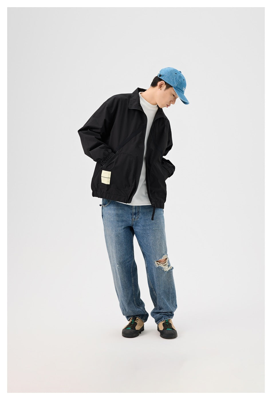 Model wearing black zip jacket over white top and light-wash jeans, hands in pockets, head tilted down