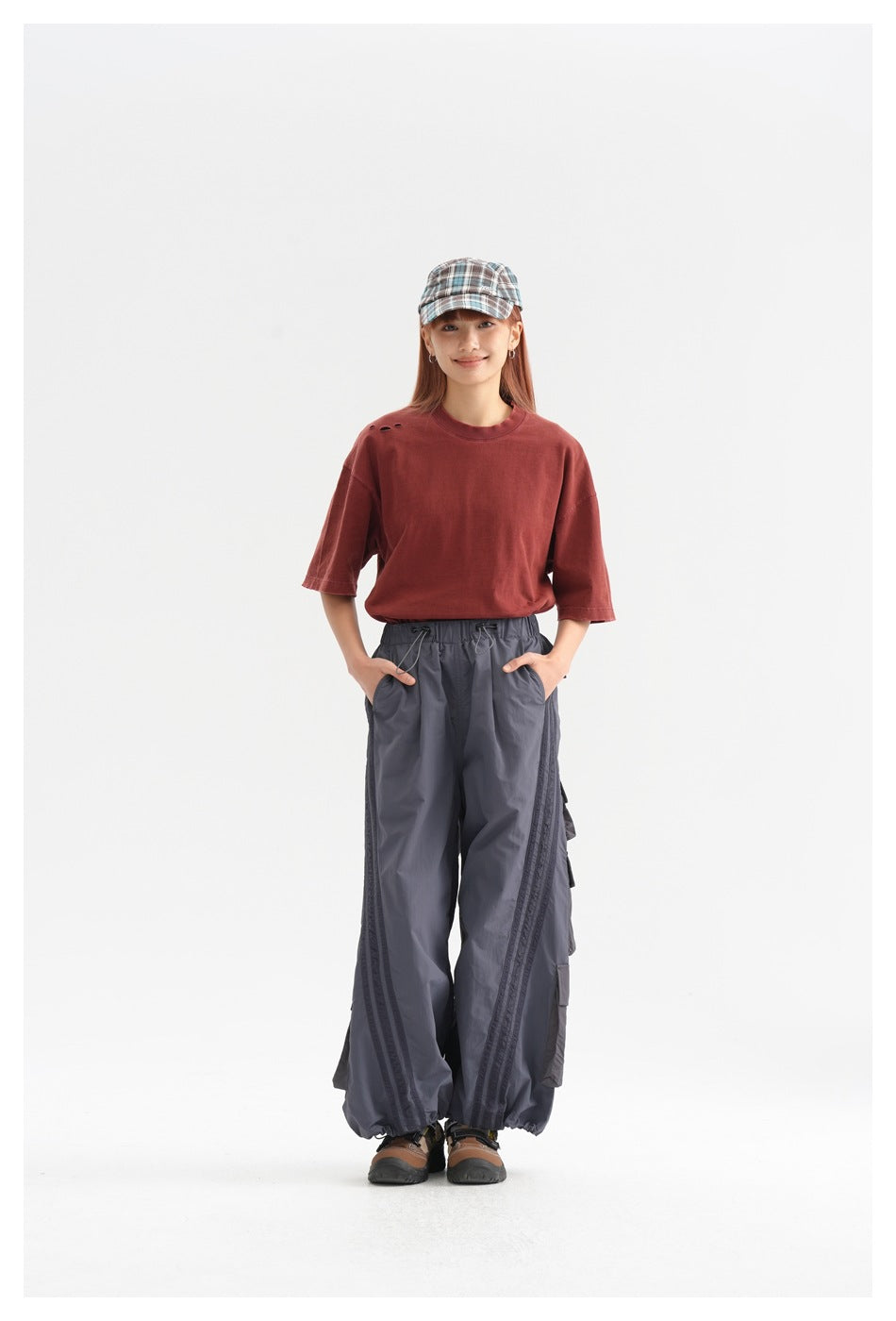 Full-length model wearing a brick-red T-shirt, loose gray cargo pants, and a plaid cap.