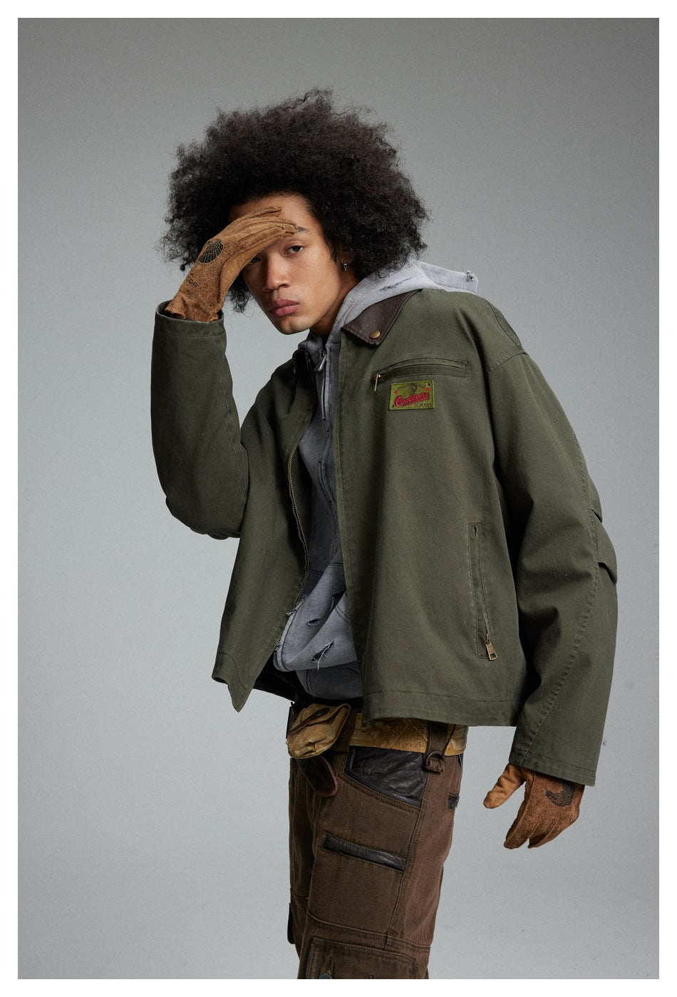 Model in an olive jacket and gray hoodie, showing the jacket's zip pockets and chest patch.