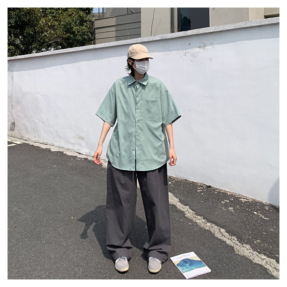 Person wearing an oversized sage-green short-sleeve button-up shirt with dark wide-leg trousers outdoors.