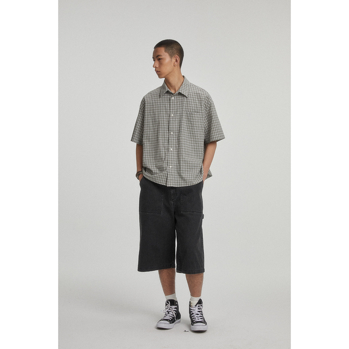 Model wearing a gray checked short-sleeve button-up shirt with a chest pocket and dark shorts.
