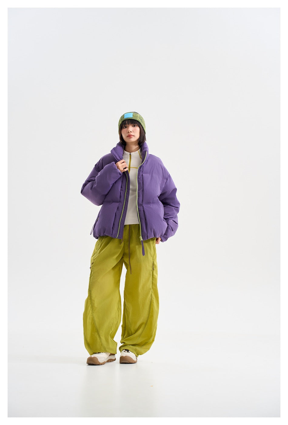 Model wearing an open purple puffer jacket over a white top, with loose lime trousers and a green knit headband.