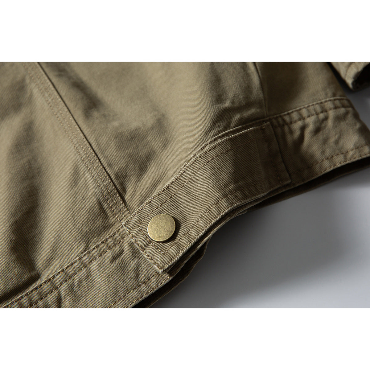 Close-up of an olive-khaki jacket hem tab with a round brass-tone snap.