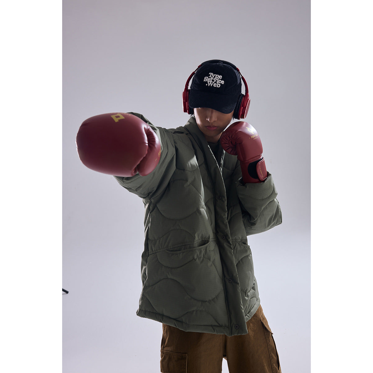 Model wearing an olive quilted jacket and boxing gloves, extending one gloved hand toward the camera.