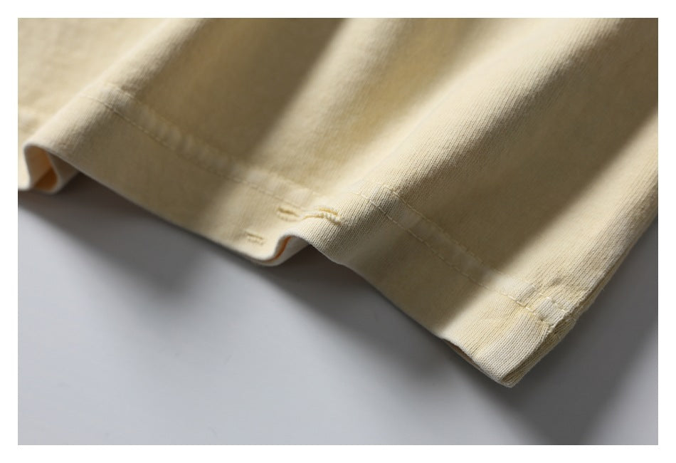 Angled close-up of the pale cream T-shirt’s folded bottom edge and stitched hem, with small distressed marks.