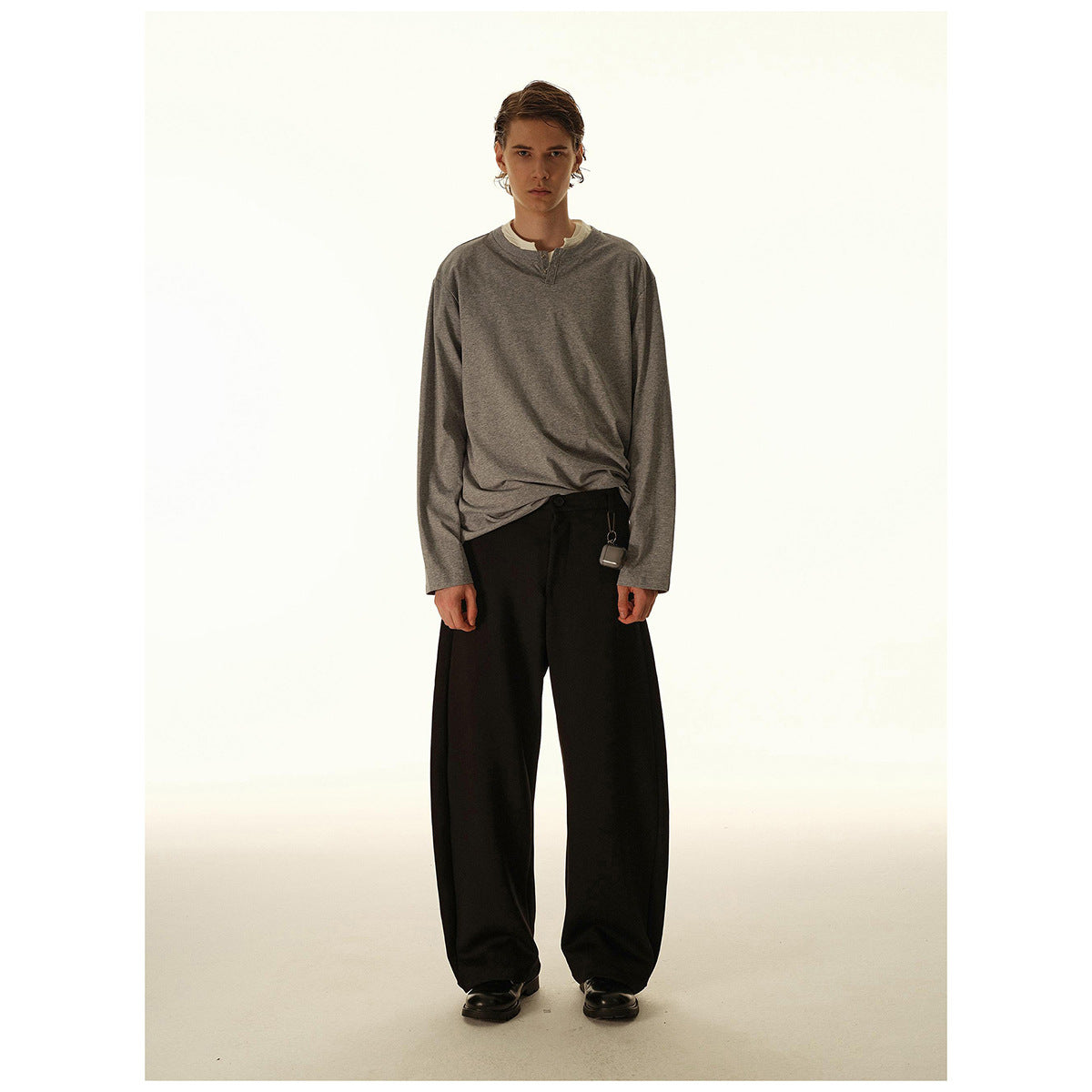 Model wearing a gray long-sleeve henley over a white layer, with loose black trousers.