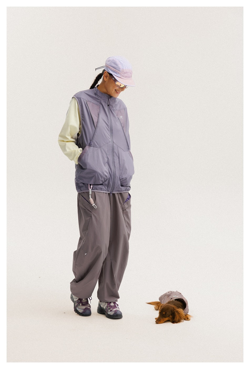 Model wearing a lavender-gray zip-front utility vest over a pale long-sleeve top, with loose gray-purple pants.