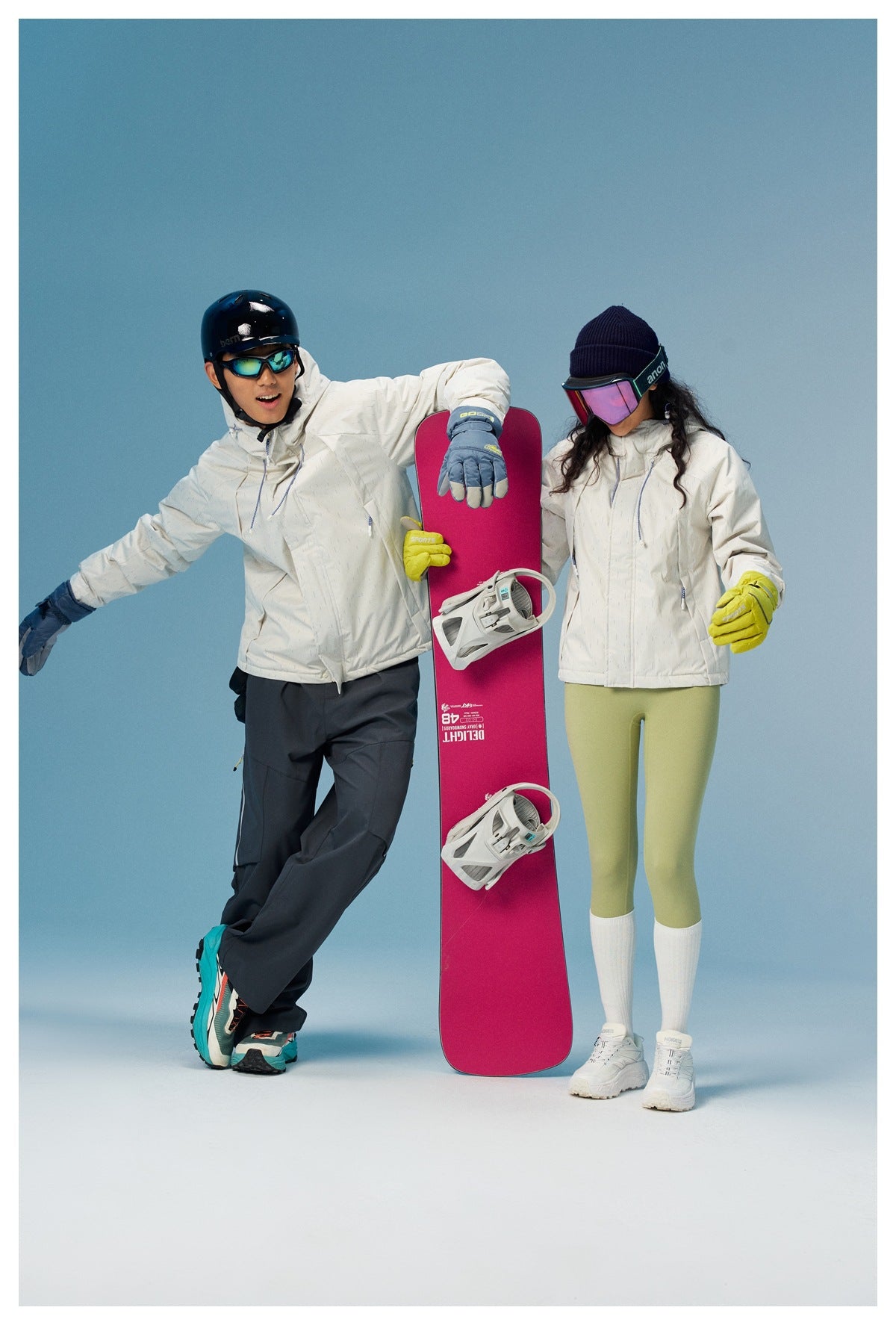 Two snowboarders stand beside a pink snowboard, wearing white jackets with dark pants and light green leggings.