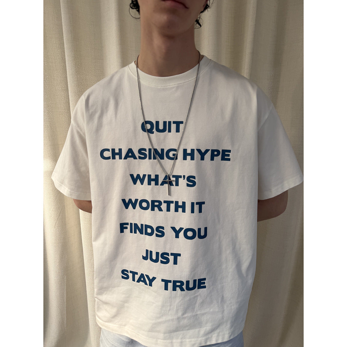 Close-up of a white T-shirt with bold blue text down the front, worn with a silver pendant necklace.