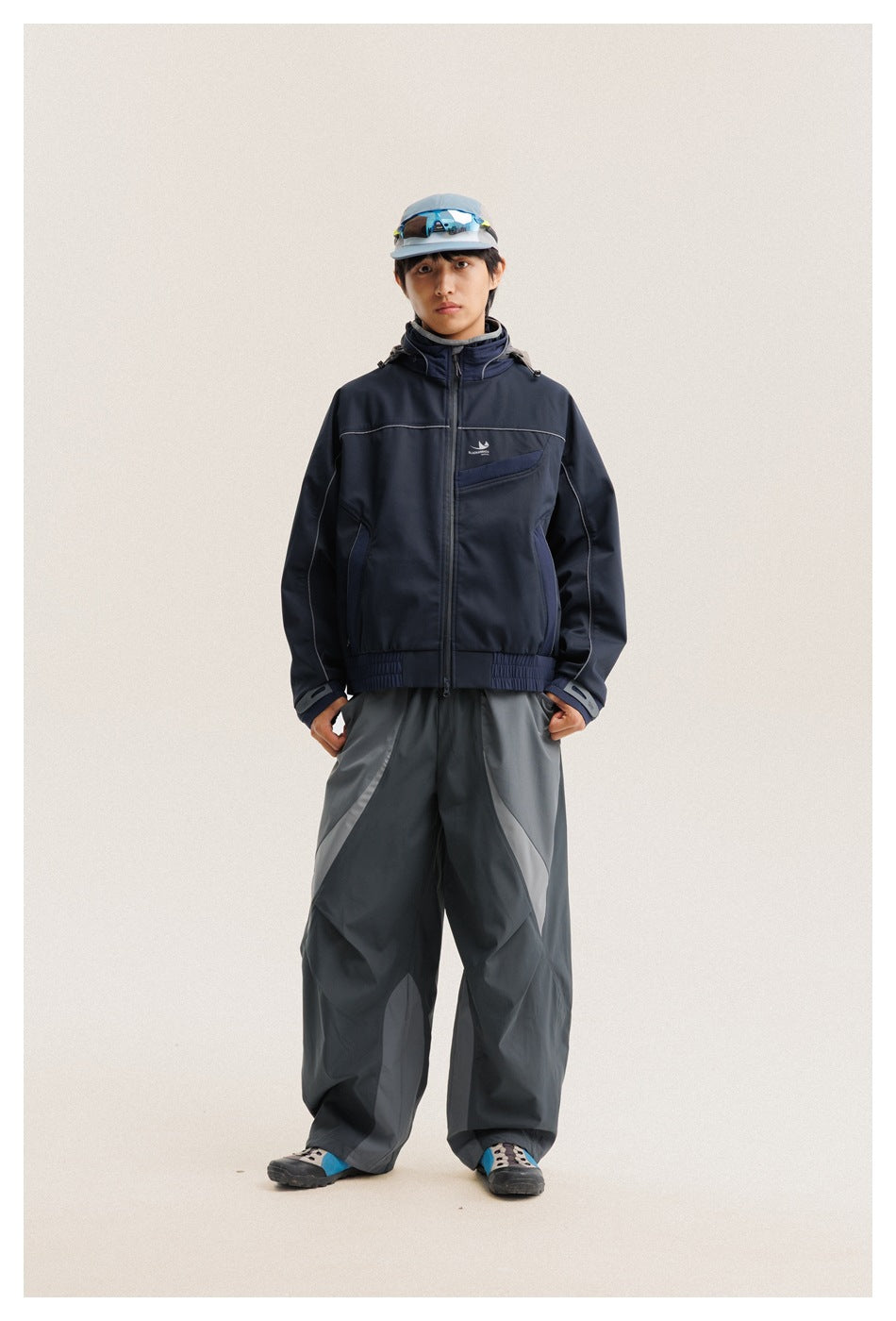 Model wearing a navy hooded zip jacket with gray piping and loose charcoal pants, shown full length from the front.