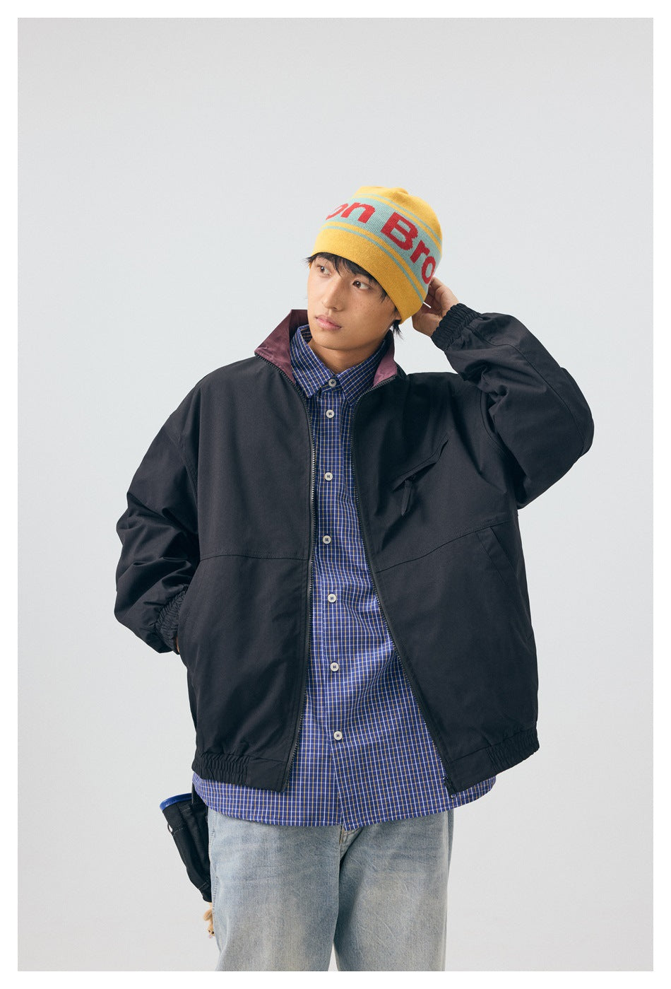 Model wearing a black zip-front jacket with plum collar lining, a blue checked shirt, and a yellow lettered beanie.