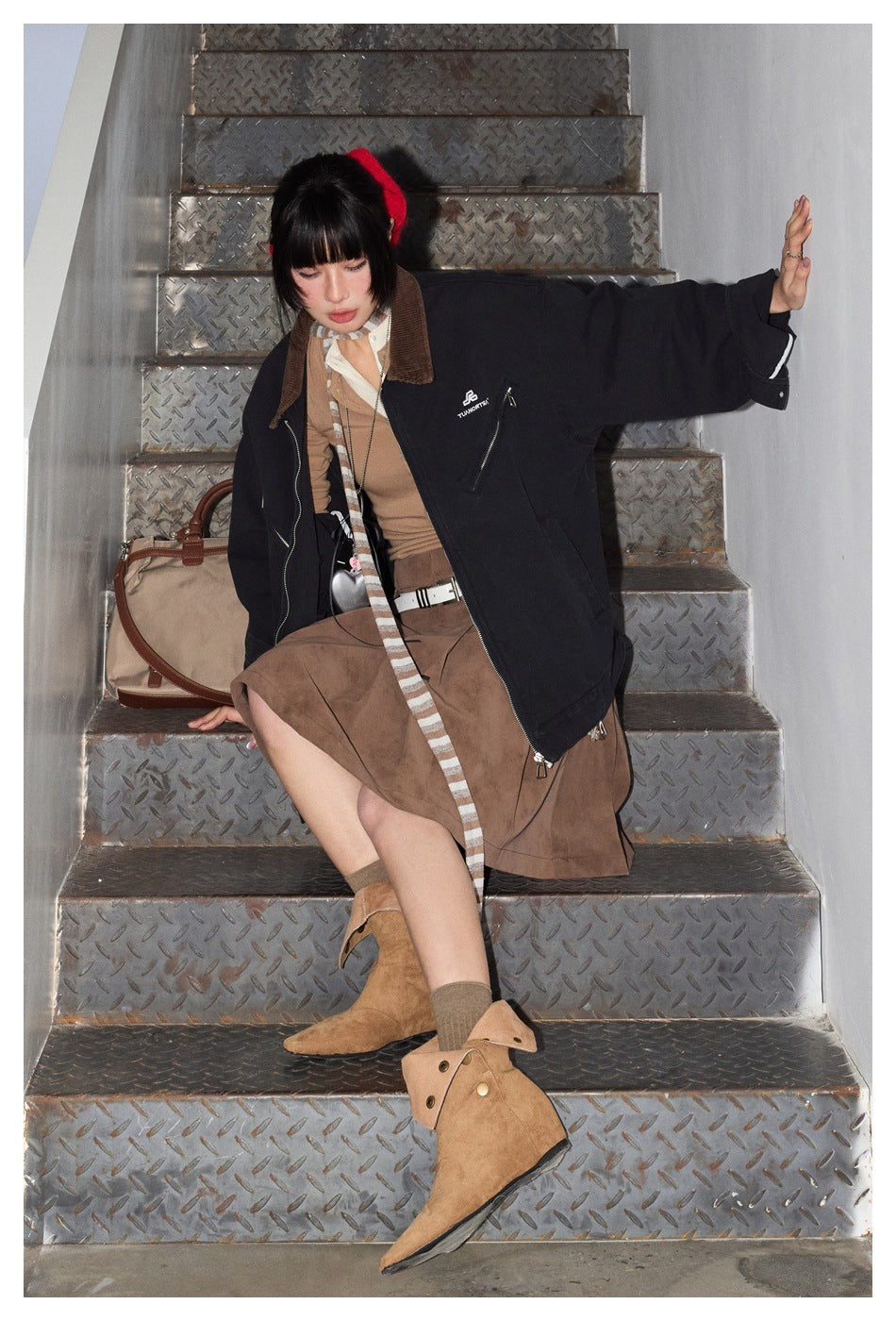 Model seated on metal stairs wearing a dark navy jacket with a brown collar and a brown skirt.