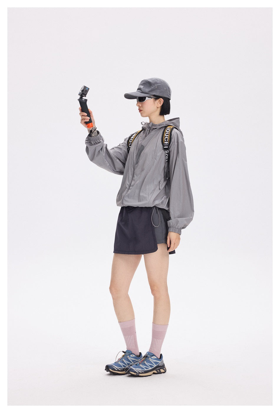 Full-length model wearing a gray hooded jacket, dark wrap skirt, gray cap, pink socks and trail shoes.