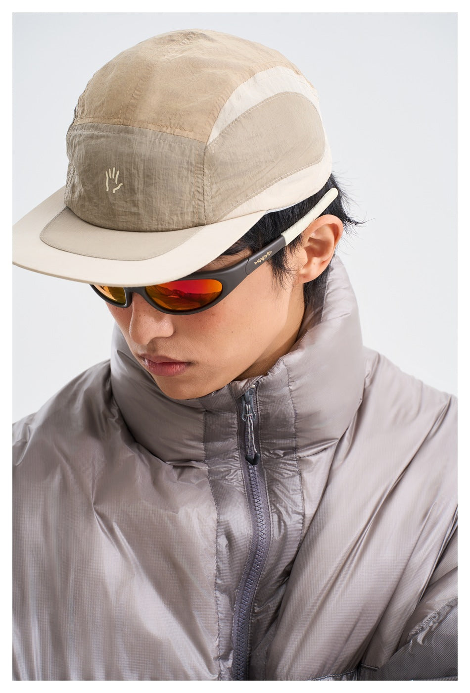 Close-up of a model in a gray puffer jacket with stand collar and zipper, wearing a cap and sunglasses