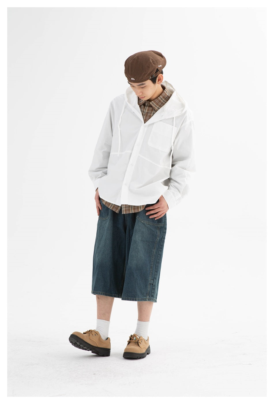 Model wearing a white hooded jacket with a chest pocket over a brown plaid shirt and wide dark denim shorts.