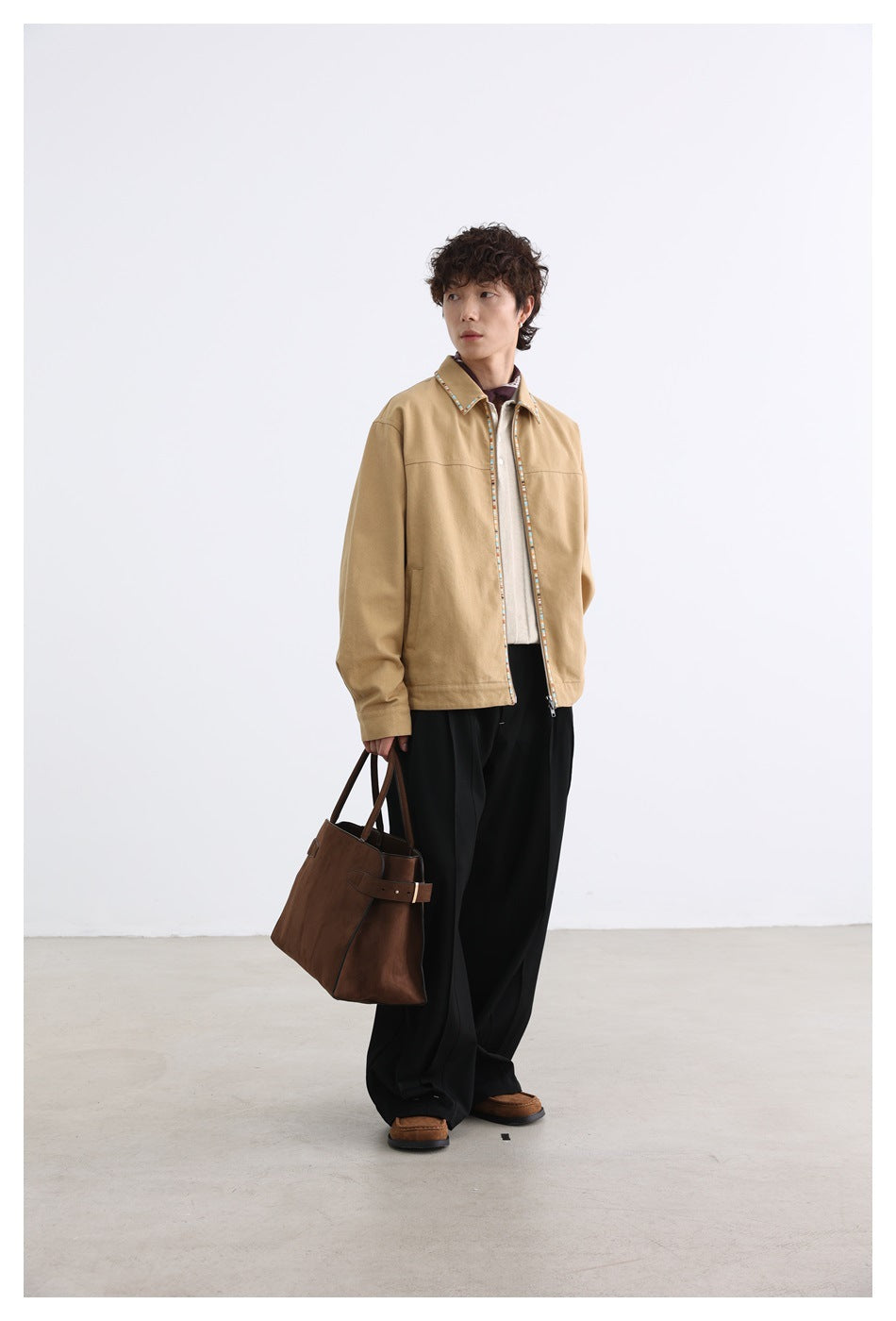 Full-length model wearing a tan jacket with multicolor trim, a light polo, black trousers, and carrying a brown tote.