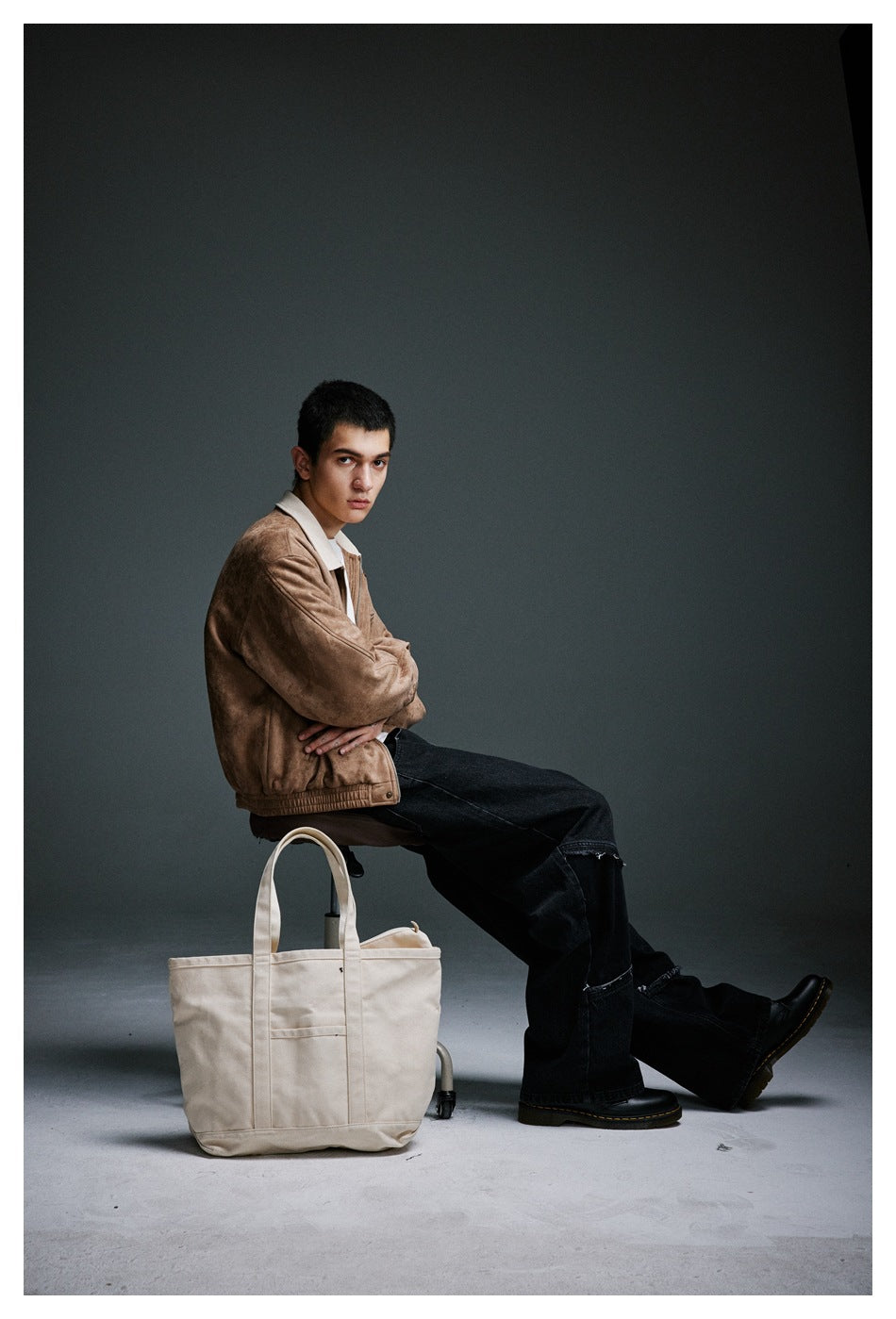 Model seated in profile wearing a tan jacket with a cream collar and black jeans.