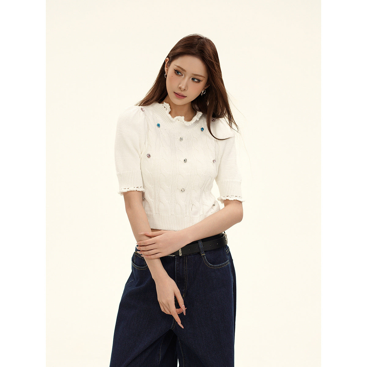 Model wearing an ivory cable-knit cropped top with jeweled accents and scalloped trim, paired with dark wide-leg jeans.