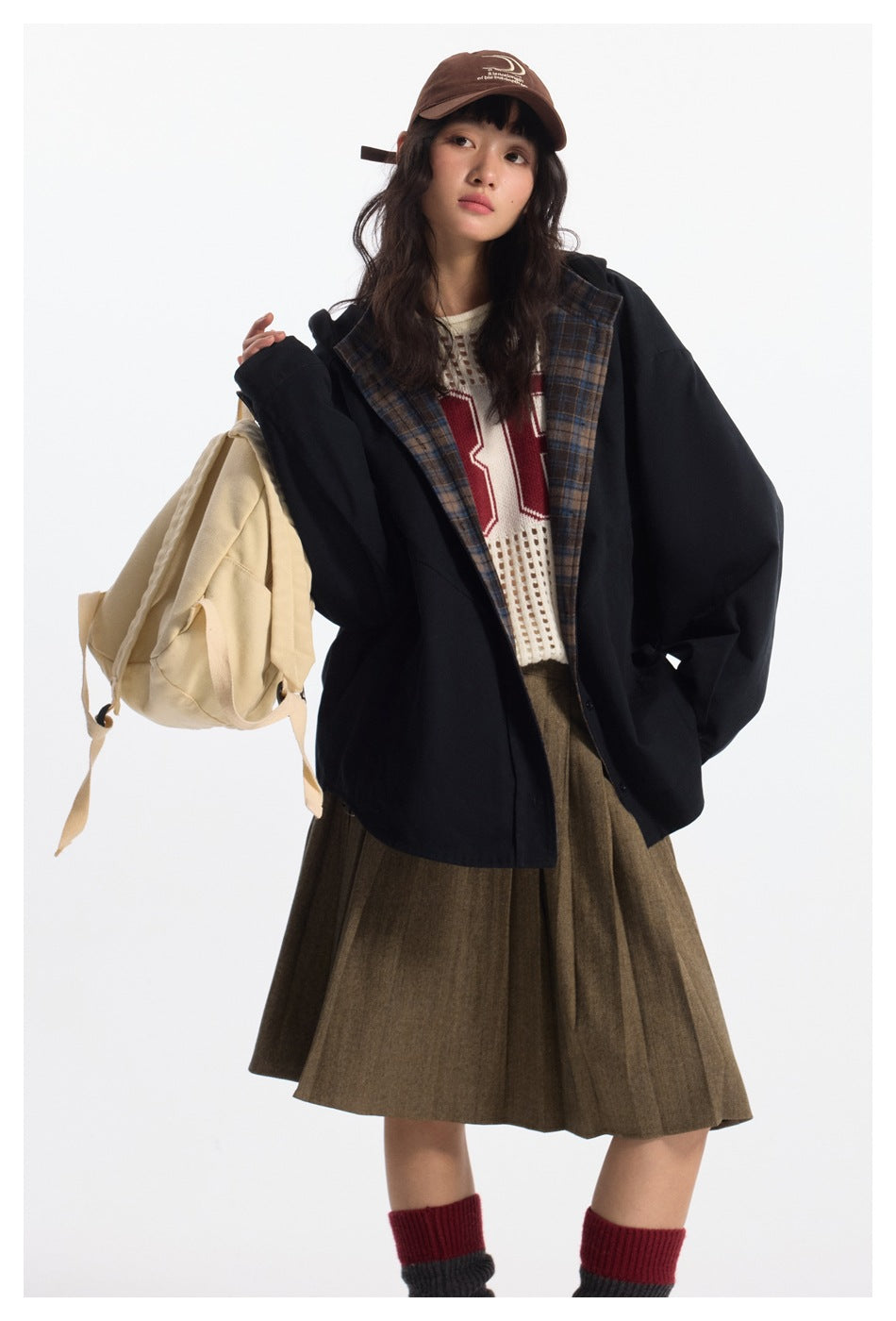 Model wearing an oversized black jacket with a brown-blue plaid lining, paired with a brown skirt and cream backpack.