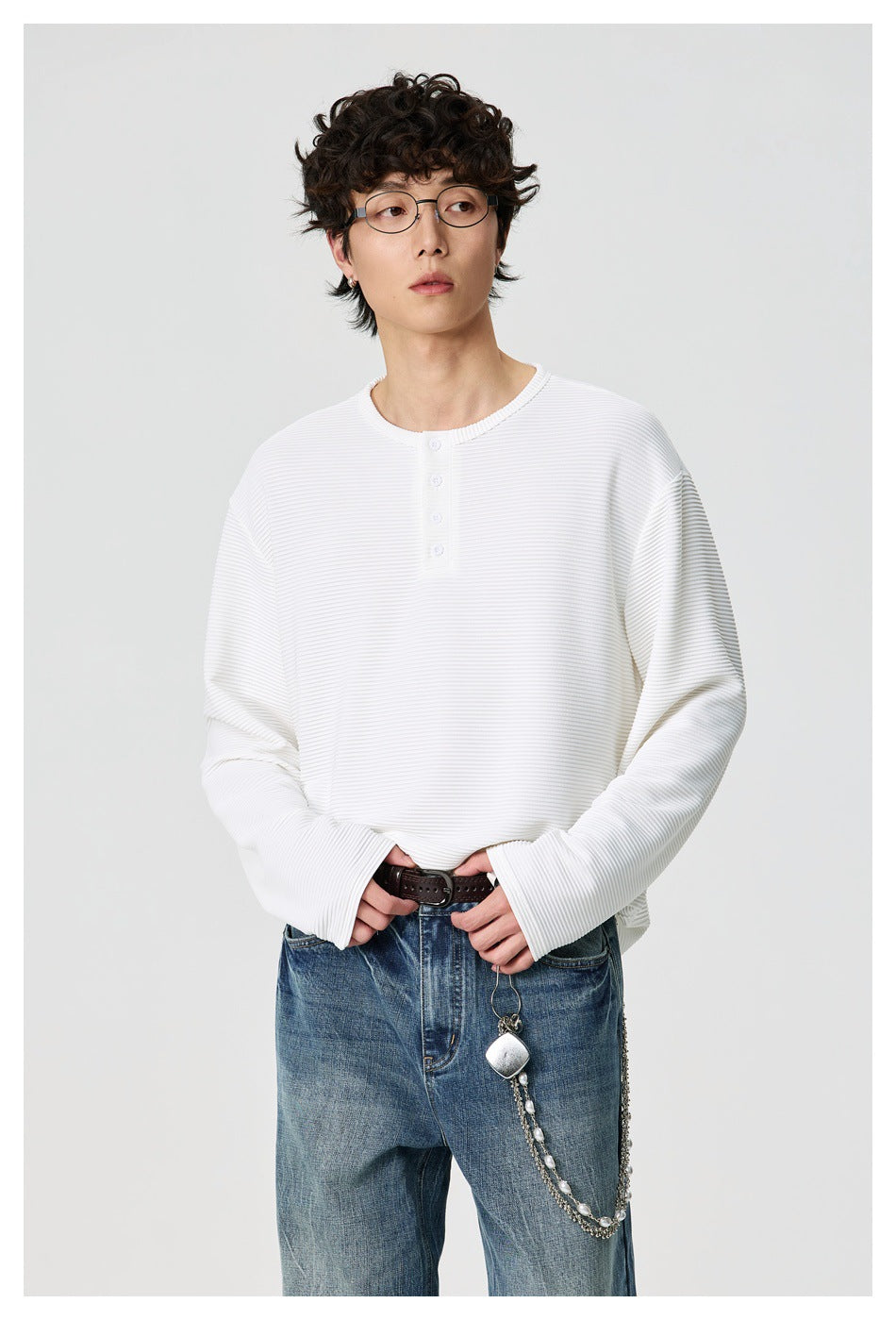 Man wearing a white ribbed relaxed long-sleeve Henley top with blue jeans, brown belt and chain accessory