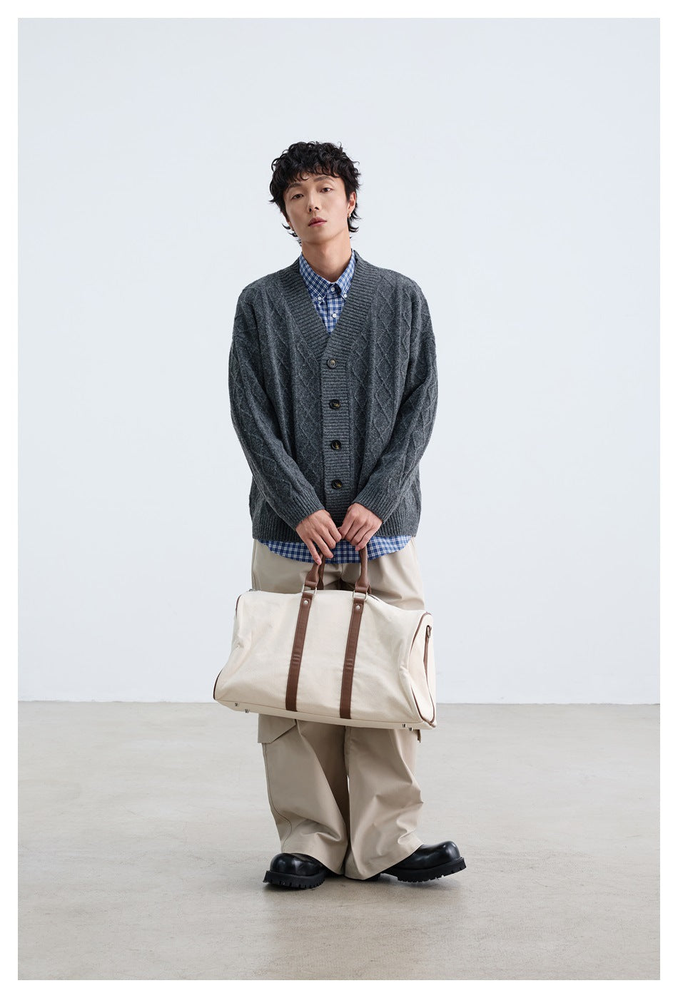 Full-length model wearing a dark charcoal cable-knit cardigan and carrying a cream duffel bag.