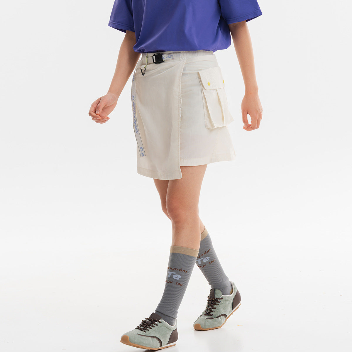 Model wearing an off-white wrap-style utility skirt with a belt, dangling strap and flap cargo pockets.