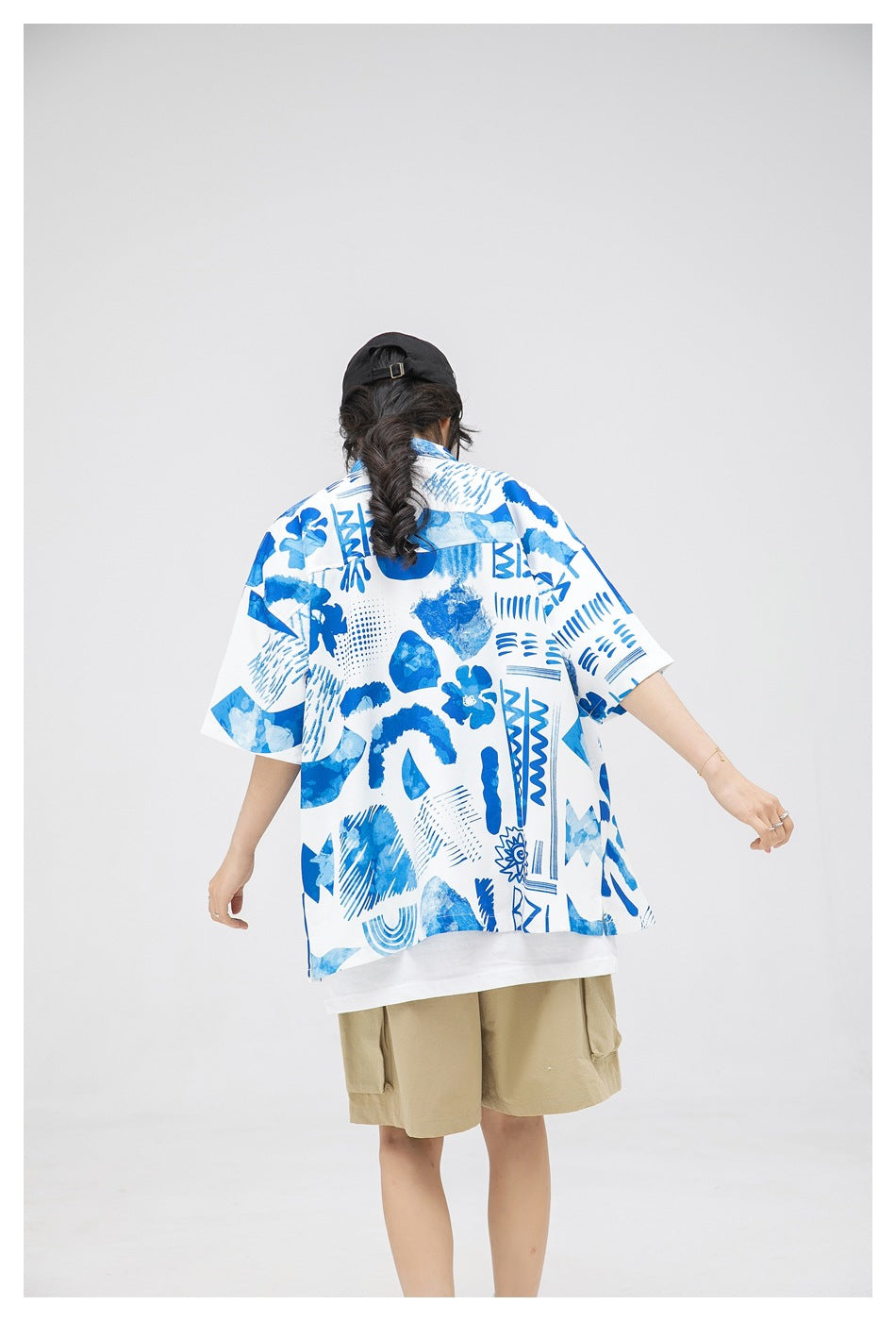 Back view of a model wearing a loose white shirt with bold blue abstract motifs and khaki shorts.