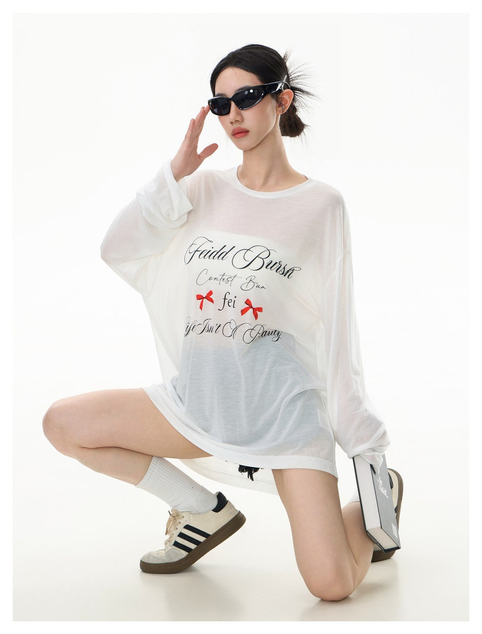 Model wearing an oversized sheer white long-sleeve tee with black script and red bow graphics, seated pose.