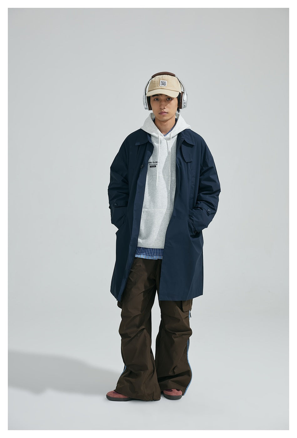 Model wearing a navy long coat open over a gray hoodie, brown cargo pants, a beige cap and headphones, front view