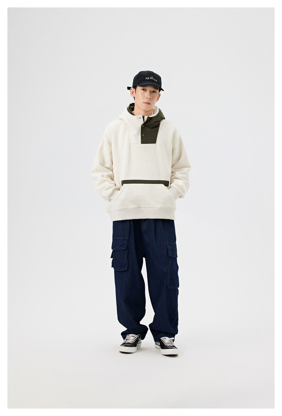 Model wearing an off-white hoodie with an olive hood and snap placket, navy cargo pants, and a black cap.