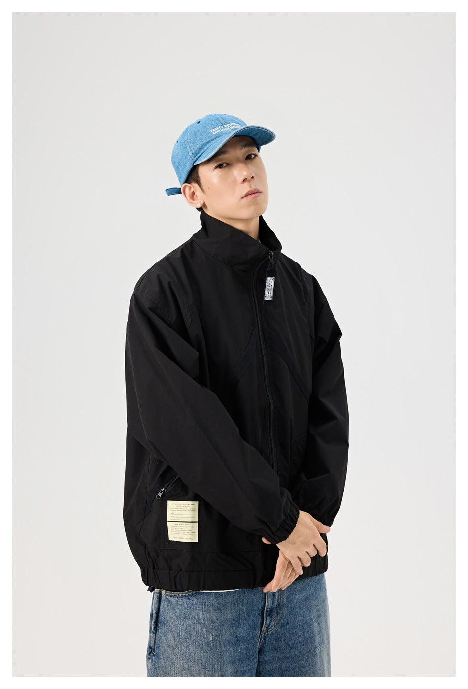 Model wearing a black zip-front jacket with elastic cuffs and hem, shown with blue jeans and a blue cap.
