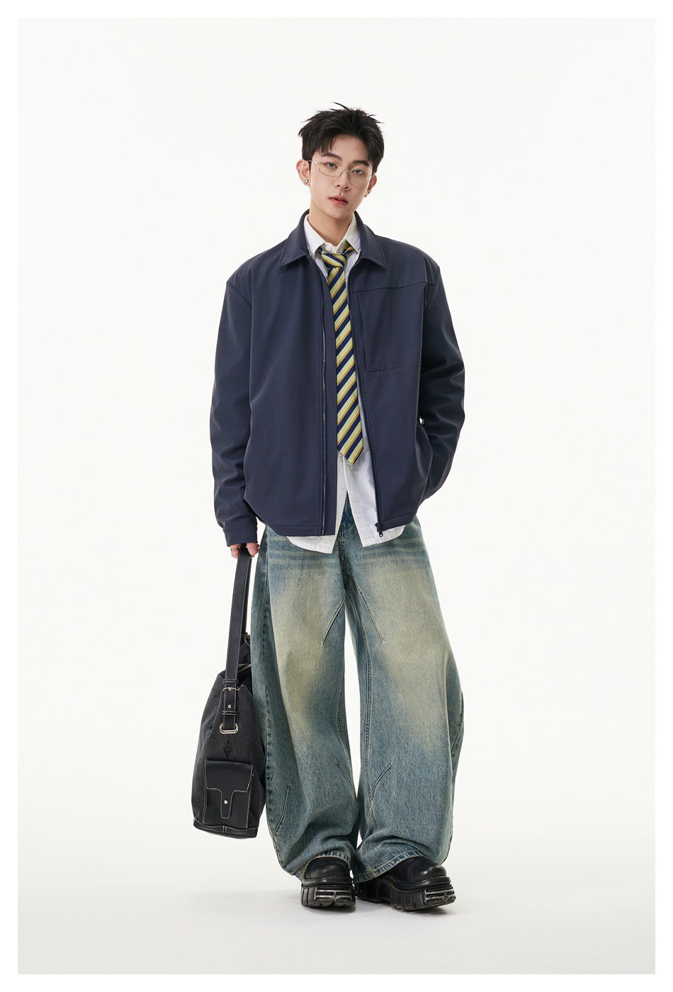 Model wearing a navy zip jacket over a white shirt and striped tie with wide-leg faded jeans, holding a black bag