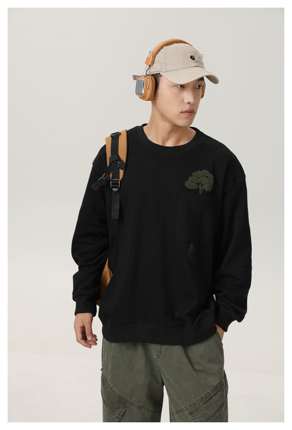 Model wearing a black crewneck sweatshirt with a chest pocket and embroidered tree, plus olive pants and a beige cap.