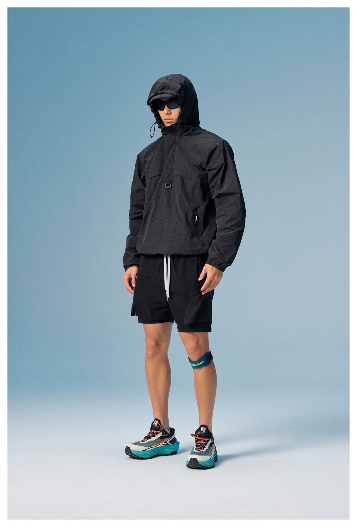 Man standing in a black hooded windbreaker and black shorts, with the hood up.