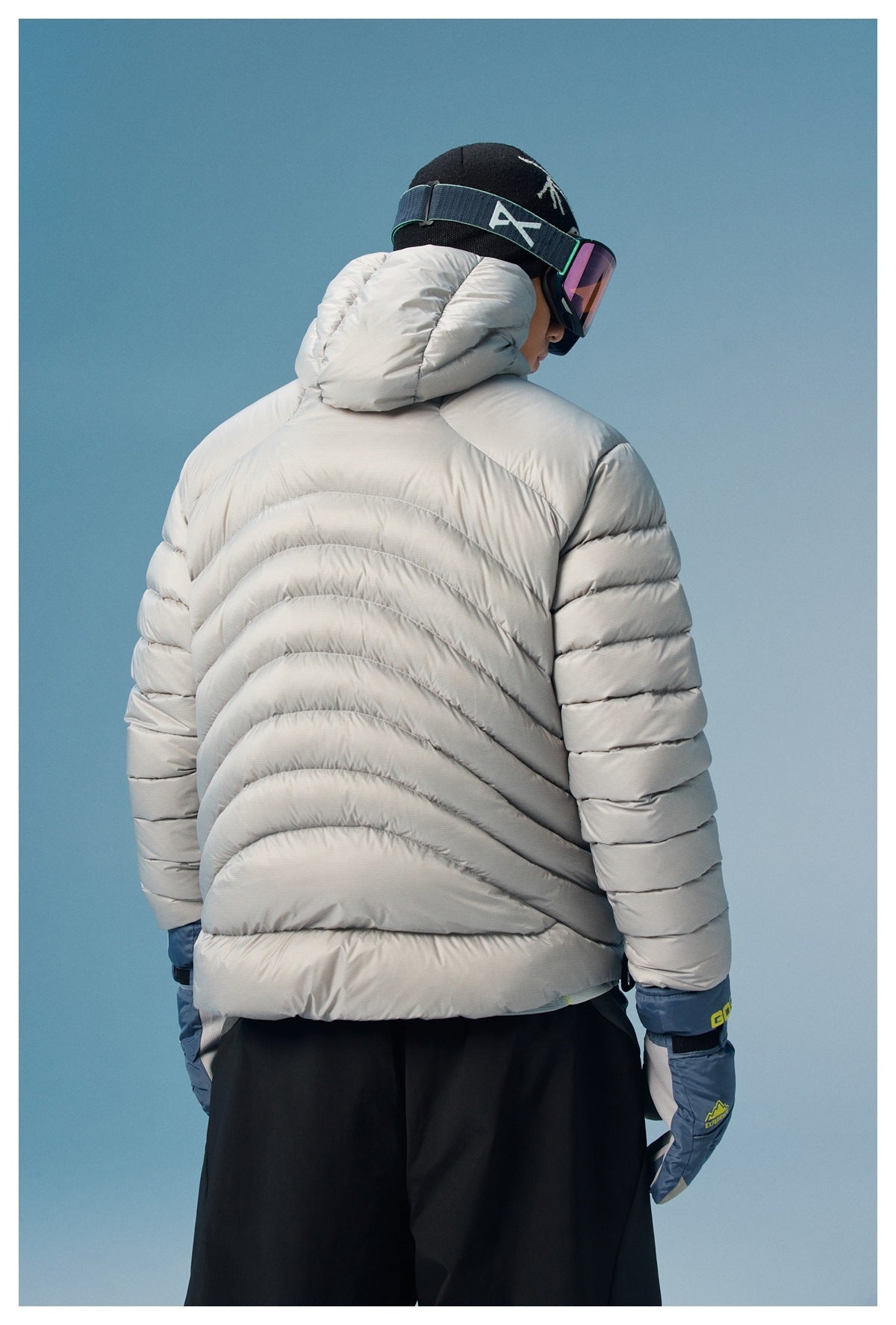 Rear view of a model wearing a light gray hooded puffer jacket and blue ski gloves.