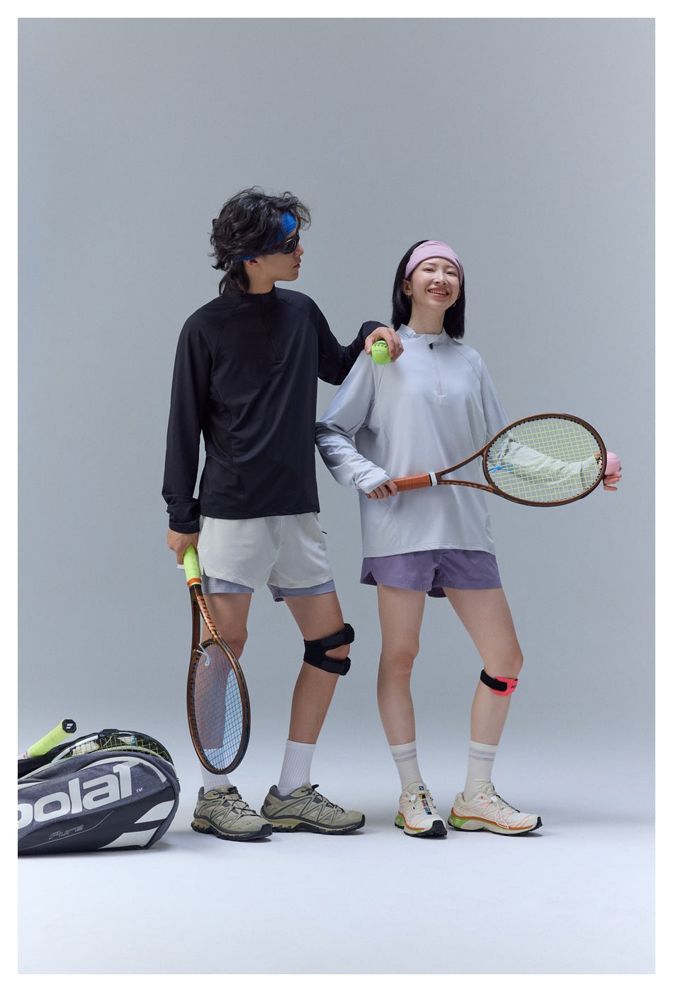 Two models pose with tennis gear; one wears a pale lavender-gray top and purple shorts, the other a black top and light