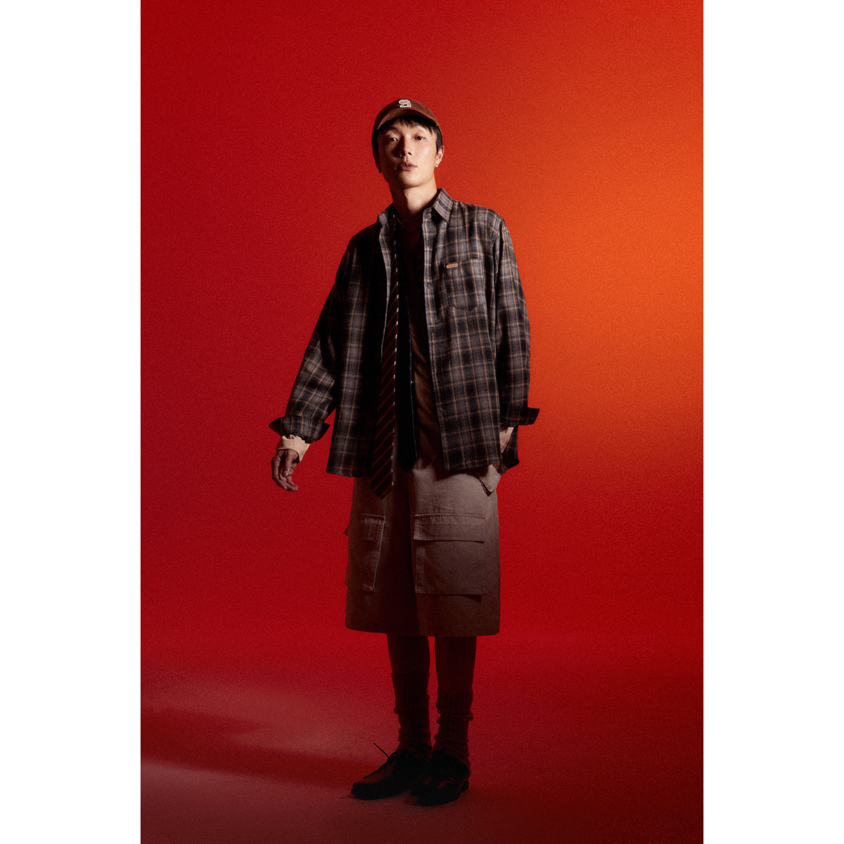 Model wearing a dark brown plaid button-up shirt over a long taupe top and cargo shorts against a red backdrop.