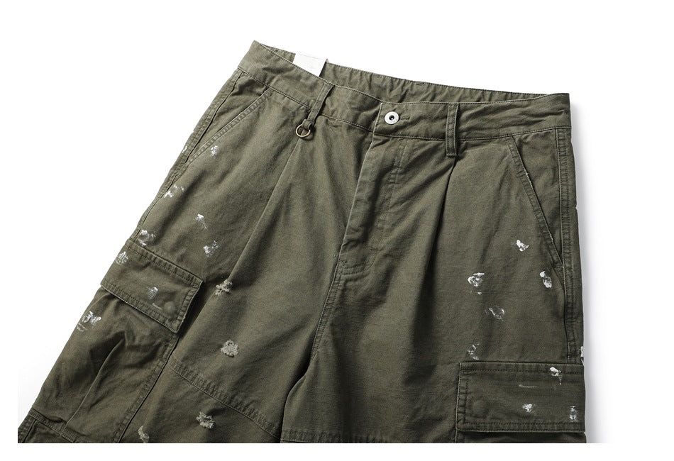 Angled front view of olive cargo pants with belt loops, a metal ring, flap pockets, and scattered white distressed marks.
