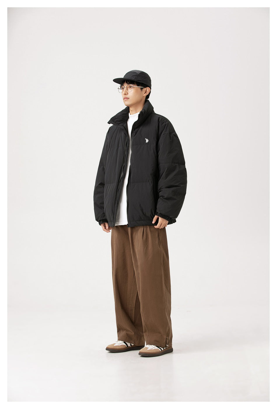 Full-length view of a person wearing a black padded jacket with a high collar, brown trousers, and a black cap.