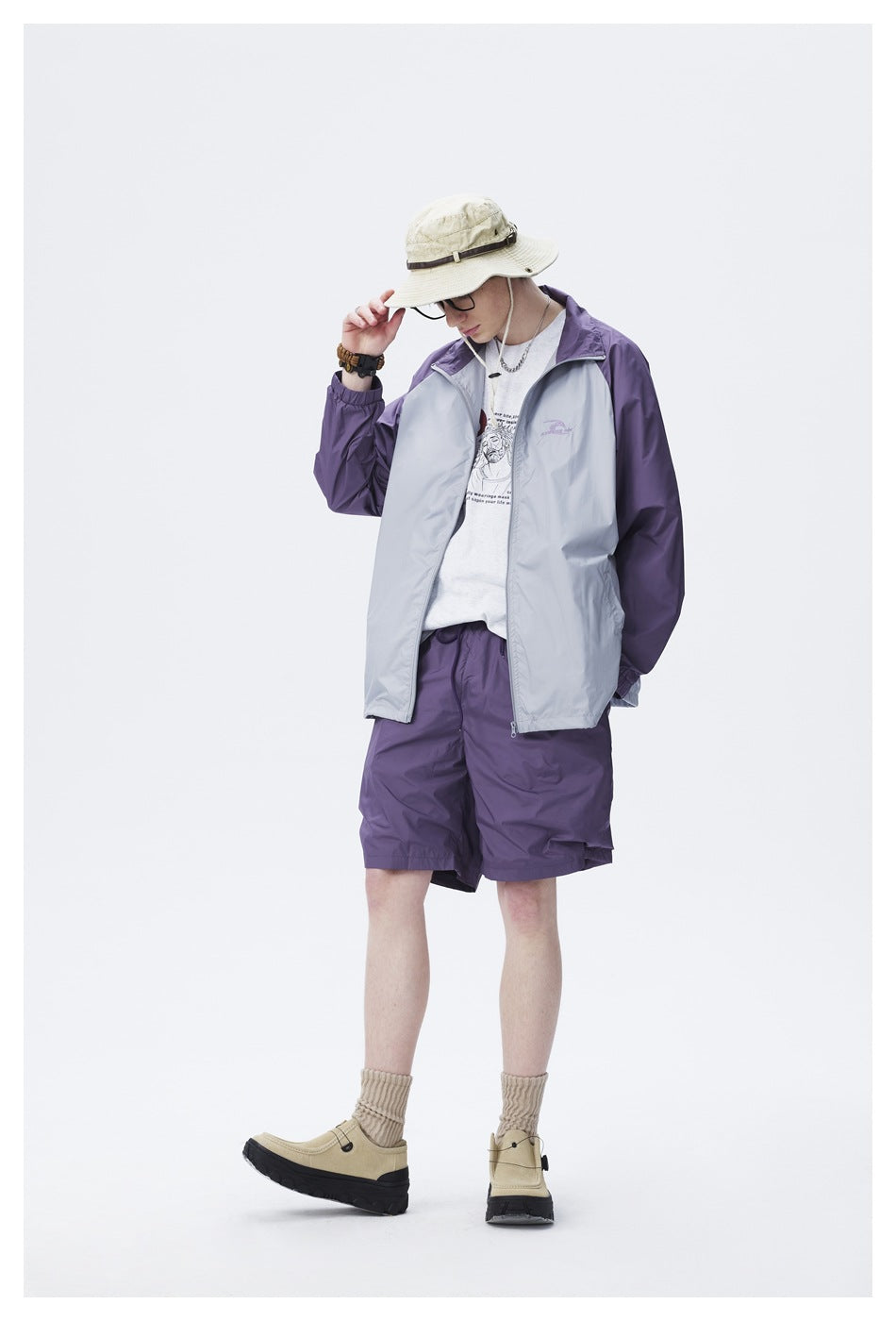Model wearing an open purple and gray color-block jacket over a white graphic tee with purple shorts and a bucket hat